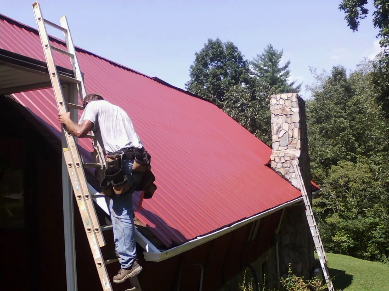 Roofer on a ladder beside a red metal roof and stone chimney — Soffit & Fascia Repair by Roofing Brothers in Farmersville